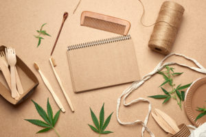open notebook with empty sheets, a textile bag and disposable tableware from brown craft paper, green hemp leaves on a wooden background. View from above, plastic rejection concept, zero waste